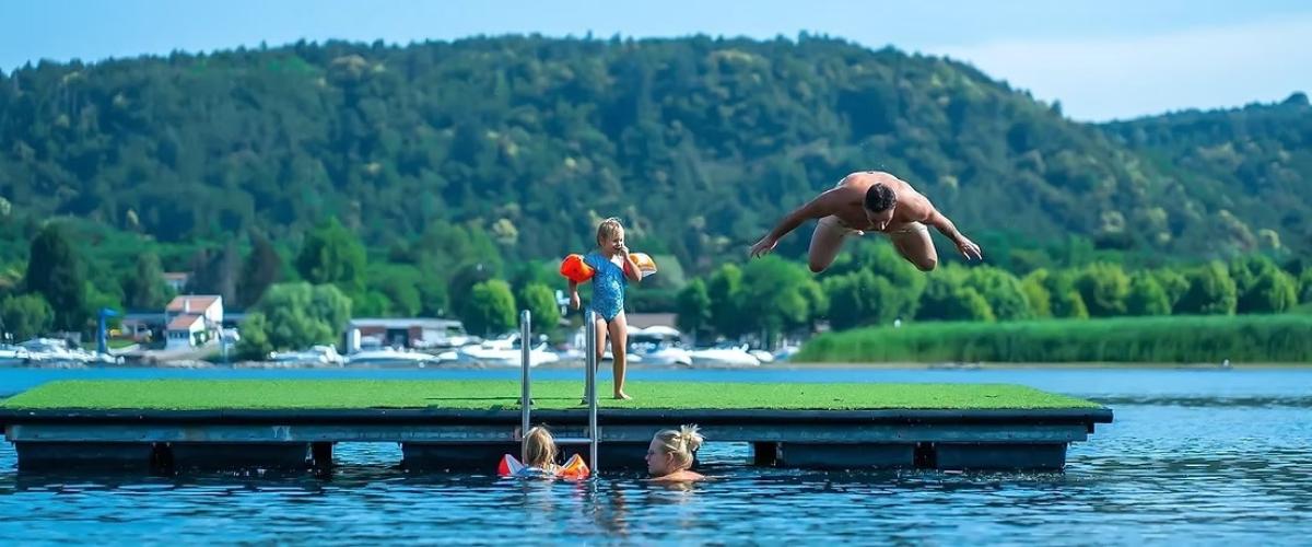 Un uomo tuffa da una piattaforma, bambini giocano felici nell'acqua, natura rigogliosa attorno.