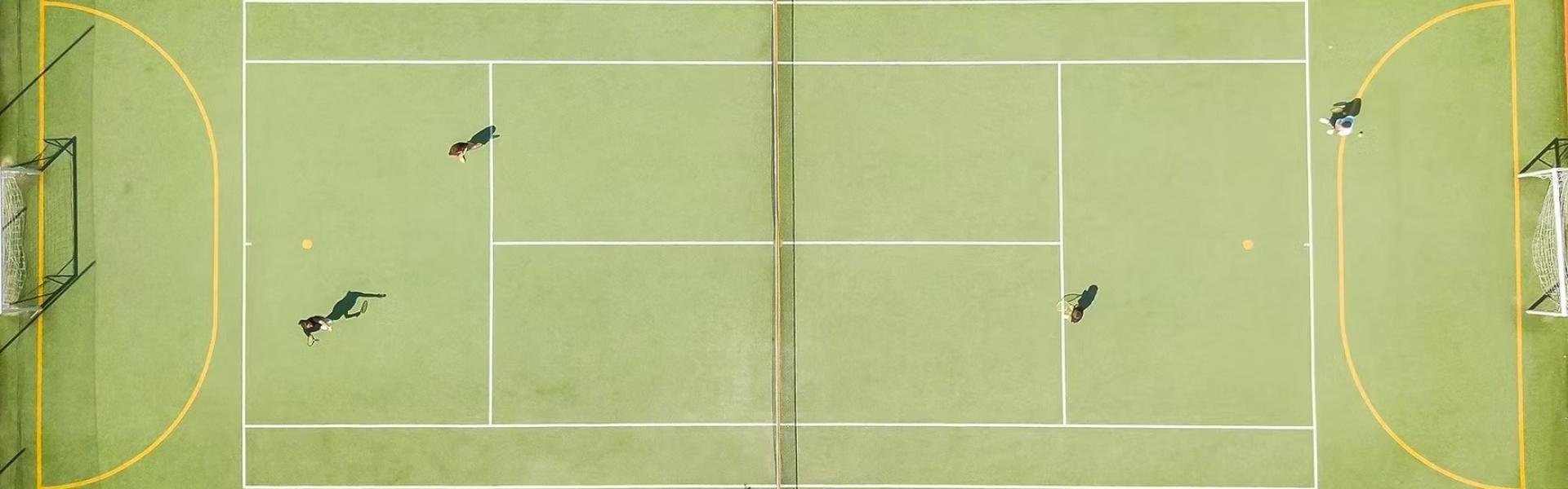 Vista dall'alto di un campo da tennis con persone che giocano, sole splendente, ombre lunghe.