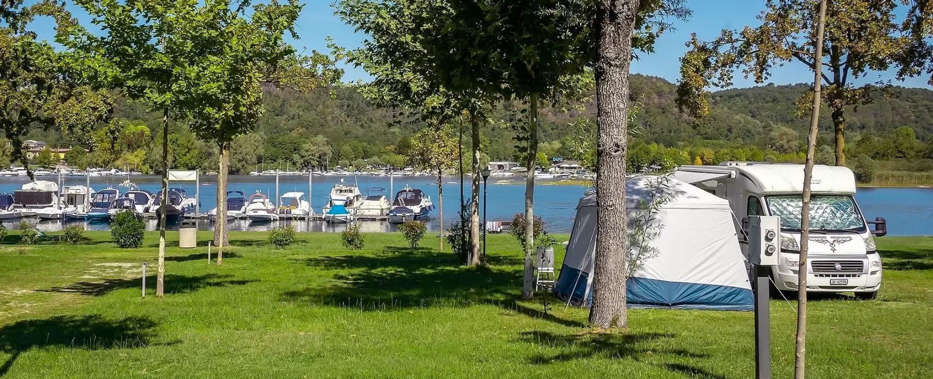 Camper e tenda vicino al lago, barche ormeggiate, natura verde, giornata serena.