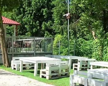 Area relax all'aperto con arredi in legno, immersa nel verde e sole splendente.