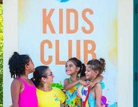 Bambini sorridenti in costumi colorati, felici davanti a un cartello 'Kids Club'.