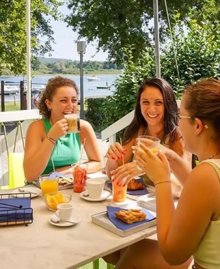 Tre amiche sorridenti brindano all'aperto, vicino al lago, godendo di una colazione estiva.