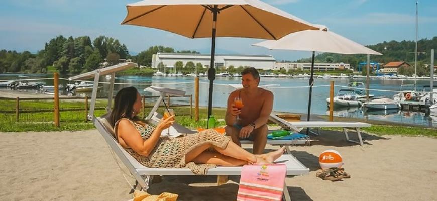 Coppia rilassata su lettini in spiaggia, sotto ombrelloni, con drink e natura attorno.