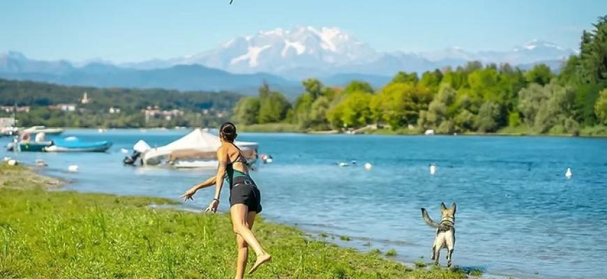 Donna gioca con cane vicino al lago, montagne innevate sullo sfondo, atmosfera serena.